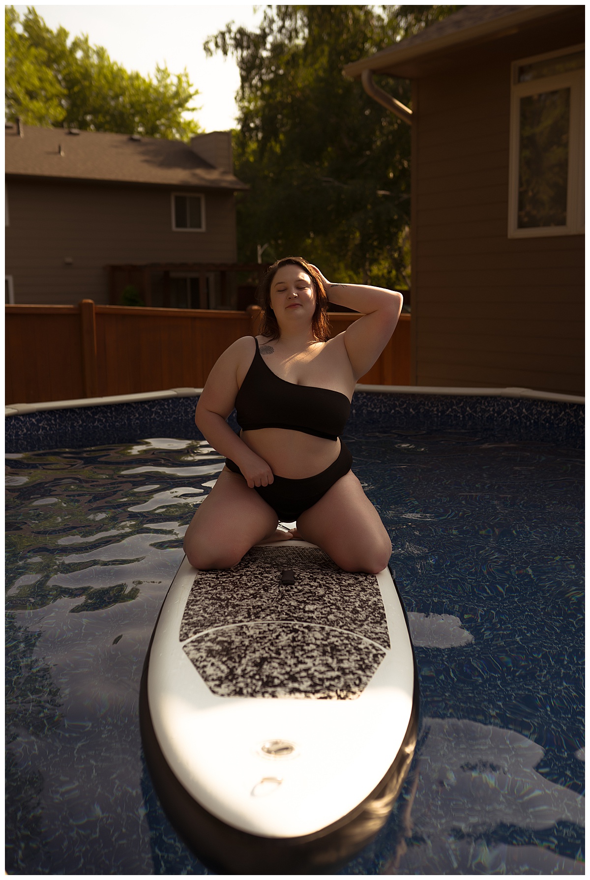 Female kneels on paddle board wearing black bathing suit during a pool boudoir session